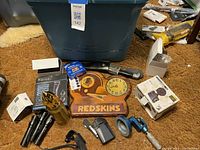 Assorted tools, gadgets, flashlights including three black flashlights, a folding knife, Redskins clock, micrometer, earplugs, and Phillips bulbs.