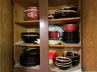Wooden cabinet shelves containing mixed bowls and plates in dark brown, reddish-brown, white, and blue colors, stacked and arranged showing quantity and variety.