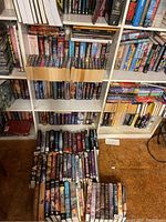 Full view of white shelving unit containing three adjustable shelves filled with mass-market paperbacks; additional books laid out on carpeted floor