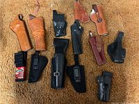Overview of various leather and nylon handgun holsters in different colors and conditions laid on carpet.