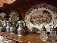 Shelf with silver-tone containers and glass salt and pepper shakers alongside a large scenic ceramic platter.