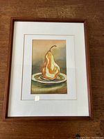 Framed watercolour artwork showing a pear on a plate with detailed brushwork and muted background tones.