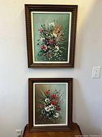 Photo showing both floral still life paintings hanging on a wall, one above the other, with brown wooden frames and green backgrounds.