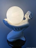 Front and side view of white ceramic Art Deco style mermaid lamp holding frosted glass globe, illuminated to show light quality.