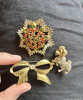 Three vintage gold-tone brooches are shown: a textured bow with a clear rhinestone, a rhinestone studded poodle, and a floral starburst medallion with orange, green, and black rhinestones.