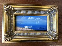 Close-up of the small oil painting showing a boat on sand with sailboats in the water, signed Y Denia, surrounded by ornate gilt frame.
