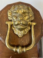 Close-up front view of solid brass devilish lion head door knocker mounted on wooden backplate, showing detailed facial features and horns.