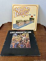 Two Max Webster vinyl LP albums side by side, showing front covers, one titled 'Live Magnetic Air' with a stage performance illustration on black background, and the other featuring cartoon heads on a conveyor belt on a cream background.