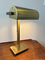 Side angle view of brushed metal banker style desk lamp on wooden surface with power cord visible.