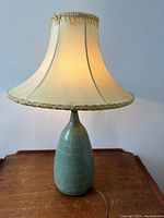 Full view of the lamp showing the green ceramic base with textured ribbing and the off-white fabric shade with fringe trim, lamp lit to show warm glow.