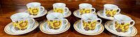 Full set of 8 matching cups and saucers arranged in a row showing yellow floral pattern and brown rims.