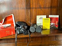 Wooden shelf with Asahi Pentax Spotmatic SP II camera in leather half-case, two cylindrical lens cases, Spotmatic manuals, and red Leitz Synchronblitzer box.
