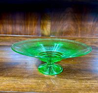 Front and side view of a bright green uranium vaseline glass pedestal compote on a wooden surface, showing its wide shallow bowl and ribbed design.