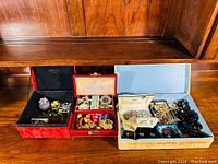 Overview of three boxes containing assorted estate jewelry pieces including brooches, rings, earrings, necklaces, and bracelets in various colors and materials.