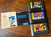 Photo of three Sega 32X cartridges stacked with visible game labels and Sonic The Hedgehog instruction manual open beside them on wooden surface.