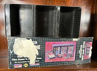 Black plastic Sega Genesis cartridge storage box with three compartments, each with vertical slots, photographed with original box behind showing wear and tear.
