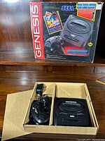 Original Sega Genesis console placed inside cardboard box compartment alongside two wired controllers and power adapter, with the original colorful box in the background.