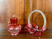 Photo of the cranberry glass pitcher and basket side by side on wooden surface with wood background