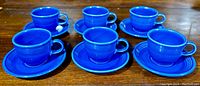 Six royal blue Fiesta Ware tea cups arranged on saucers on a wooden surface, viewed from the front revealing shape, color, and design.