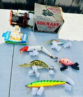 Photo showing six vintage fishing lures arranged on a white surface, including a packaged Kwikfish lure and a Normann No. 29 spinning reel with its original box.