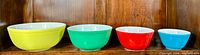 Four vintage Pyrex mixing bowls in primary colors (yellow, green, red, blue) arranged side by side showing size differences and colors.