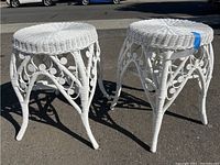 Pair of white wicker side tables/plant stands shown together outdoors on asphalt for size and condition view. Both have round tops and curved legs with scroll pattern.