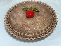 Top view of ceramic pie keeper lid showing red apple handle and textured brown surface.