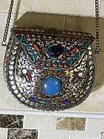 Front view of the semi-circular clutch bag showcasing detailed etched metal design in silver and gold tones, decorated with colorful stones mainly blue, red, turquoise.