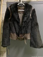 Front view of black fur and leather jacket hanging on a hanger showing overall construction, fur material, and leather belt.