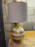 Full view of the Kate Spade New York table lamp showing round base with gold and white stripes and beige fabric shade off.