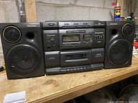 Front view of Panasonic stereo system with two black detachable speakers on a wooden surface, showing cassette deck with buttons and dials, radio tuner display and CD player lid on top.