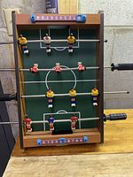 Front view of table football game showing red and yellow player figures, white football field markings, wood frame, and manual sliding scoreboards on top and bottom ends.