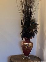 Full view of red glass vase with gold leaf and decorative branches with peacock feathers inside.