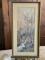 Front view of vintage signed artwork depicting cabin by creek among birch trees, framed vertically with goldtone frame and cream matte.