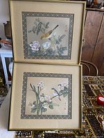 Two framed Asian-style watercolor paintings on silk, each depicting a bird and floral elements, signed by the artist, shown stacked.