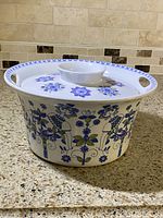 Side view of the white porcelain casserole with blue and green floral and girl patterns, showing the lid with handle and wall sockets.