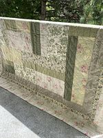 Full view of the quilt hanging outdoors showing patchwork design with various floral and leafy fabric panels in pastel greens, pinks, and beige tones.