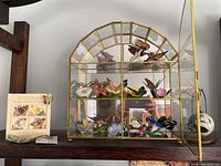 Full frontal image of the brass and glass display case housing the butterfly figurines with the butterfly book and pin displayed on the left side.
