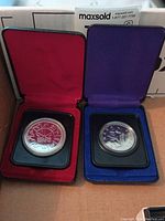 Two silver commemorative coins inside velvet-lined cases; one red-lined, one purple-lined, each coin in a protective holder.