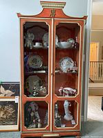 Full front view of painted Asian style display case showing glass front doors and interior glass shelves with collectibles displayed.