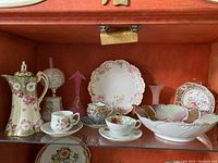 Wide view showing collection of China and glass items including Nippon teacups, coffee pot, cranberry glass bell, pink glass vase, and hand-painted oil lamp.