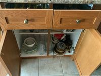 Open wooden cabinet with white and green patterned plates stacked on left side, clear glass drinking glasses in front, and various white ceramic bowls and ramekins stacked on right side along with kitchen utensils.