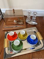 Photo showing covered cheeseboard with clear dome lid and wooden base, cheese cutting utensils, stainless steel tray with gold coloured handles, and four colorful espresso cups on saucers arranged on the tray.