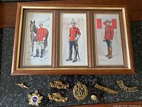 Framed trio of embroidered RCMP military art featuring officers in red lined coats with horse and Canadian flag, placed above assorted brass military pins and badges on black stone surface.