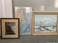 Three nautical-themed pieces displayed on a floor: a black and white Bluenose II print, a Ghost Fleet map, and a large lighthouse painting.