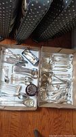 Two boxes filled with assorted silver plated flatware including spoons, forks, ladles, and serving utensils displayed on a wood floor.