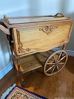View of wood tea cart showing carved wood details on side, glass tray on top, large wooden spoked wheels, and wooden shelves below.