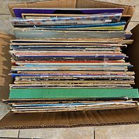 Top-down view into a cardboard box containing a large stack of vinyl records showing many album spine edges.