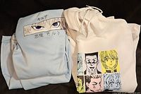 Two anime hoodies folded and displayed. One light blue with a blonde-haired character and Japanese text, the other white with six Hunter x Hunter characters in colored squares.