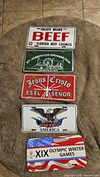 Photo of five assorted decorative and themed license plates with various slogans and designs including Florida Beef Council, St. Joseph Co. Grange Fair, a religious plate, America eagle, and Olympic Winter Games.